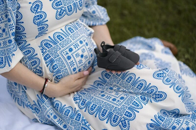 Mulher grávida com vestido estampado azul e branco, segurando sapatinhos de bebê cinzas na barriga