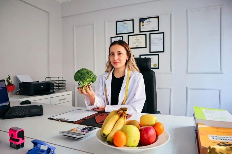 Nutricionista sorrindo em consultório, segurando um brócolis, com frutas na mesa