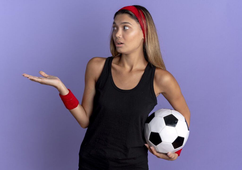 Jovem garota fitness em roupas esportivas pretas e bandana vermelha, segurando uma bola de futebol